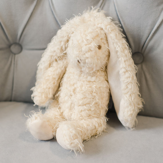 A Grace Bunny with fluffy white fur and an embroidered face, sitting on a gray tufted sofa. The bunny appears well-loved and scruffy, perfect as a baby gift.