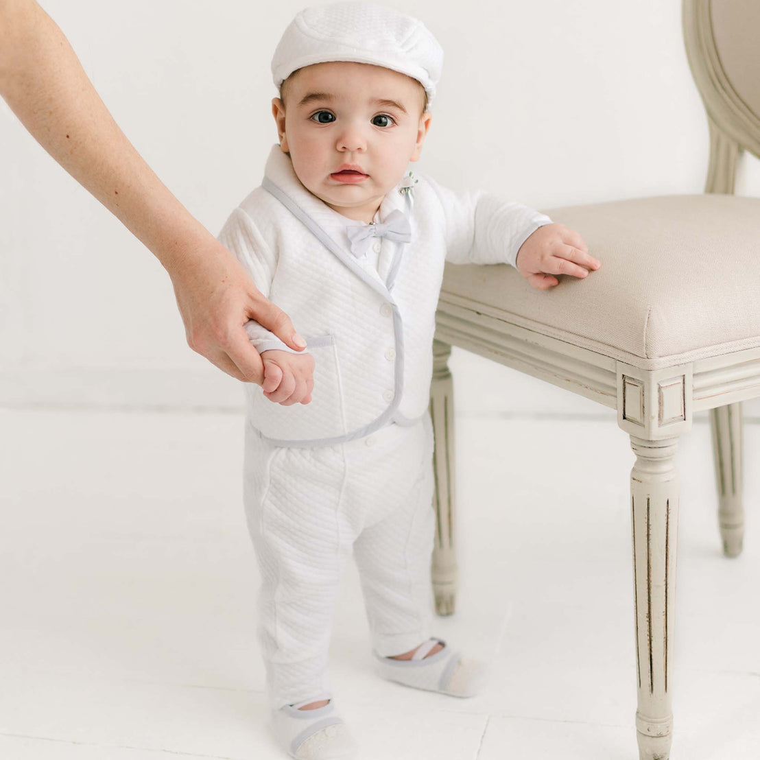 A baby dressed in a Harrison 3-Piece Pants Suit and cap stands while holding an adult's hand next to an elegant chair in a light-filled room.