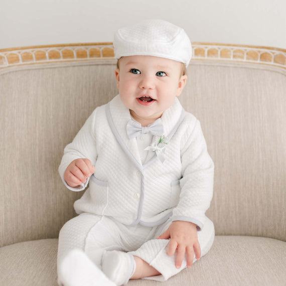 A baby in a white Harrison 3-Piece Suit and cap sits smiling on an upscale beige sofa, holding a small pose.