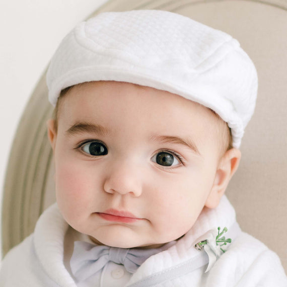 A close-up portrait of an adorable baby with big eyes and a slight smile, wearing a Harrison Textured Newsboy Cap in white and a white outfit with a collar.