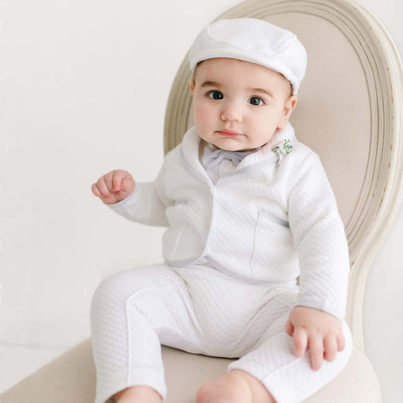 A baby boy wearing a stylish white Harrison 3-Piece Pants Suit and matching cap, sitting on an elegant cream chair against a light background. The baby looks curious and thoughtful.