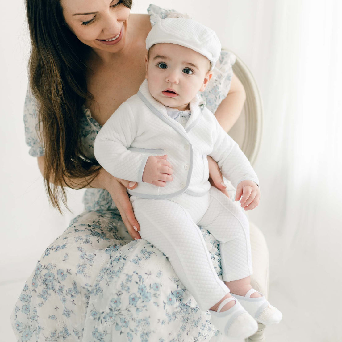 A smiling woman in a floral dress holds a baby dressed in a Harrison 3-Piece Pants Suit, sitting in a bright, airy room. The baby, wearing a white hat, looks curiously at the camera.