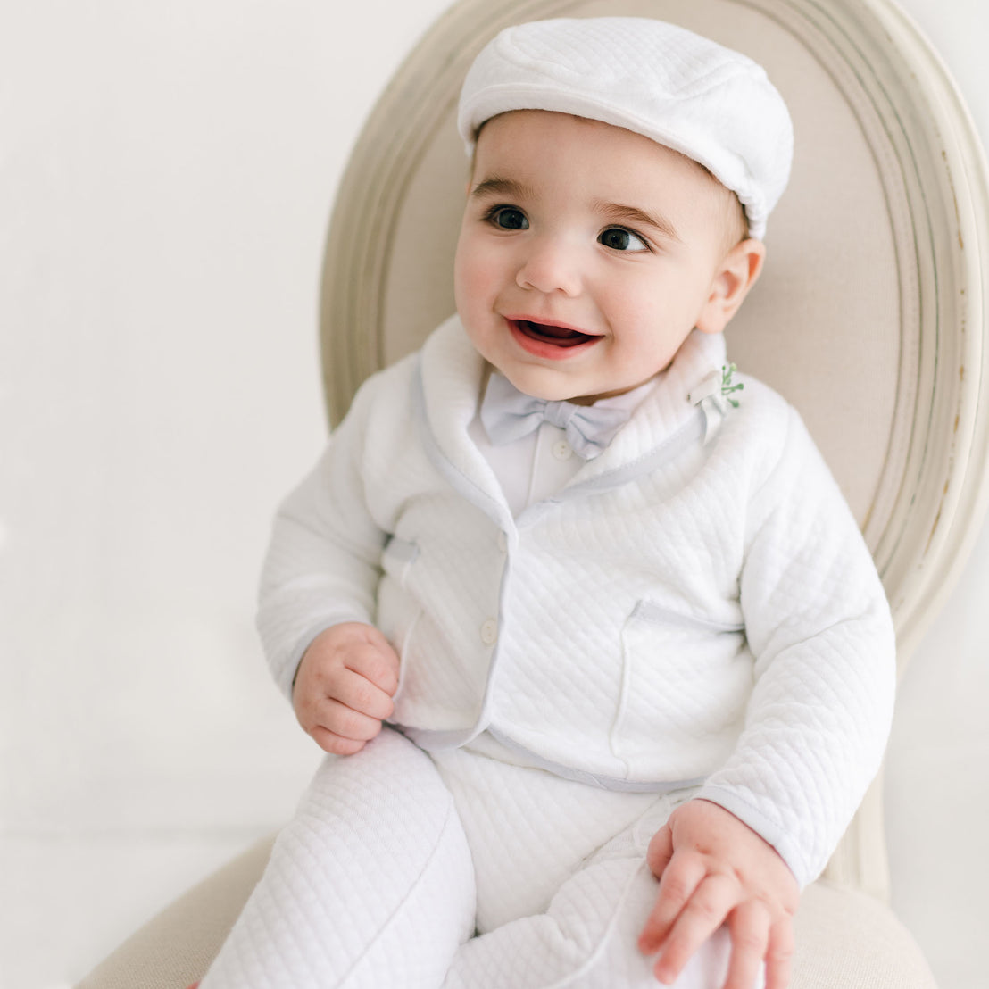 A smiling baby dressed in a Harrison 3-Piece Pants Suit and cap, sitting on an antique-style chair with a plain background, exuding happiness.