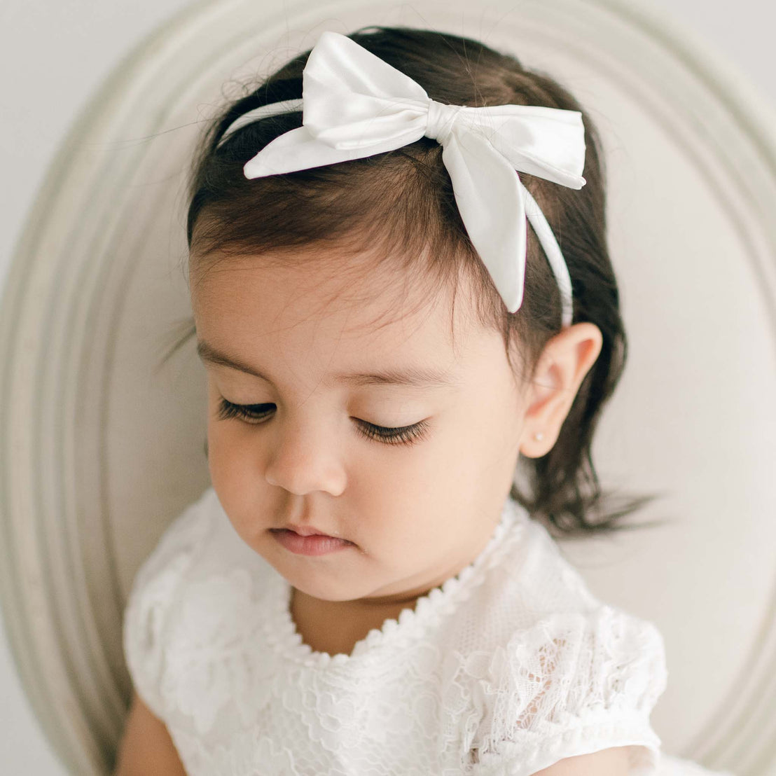 Baby girl with dark hair wearing the Victoria bow headband in Ivory. 