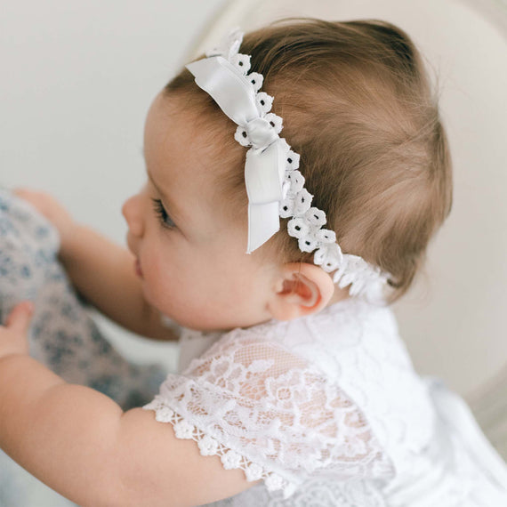 A close-up of a baby girl wearing a delicate white lace dress and the Olivia Headband with a bow, suitable for her christening or baptism, looking gently to the side.
