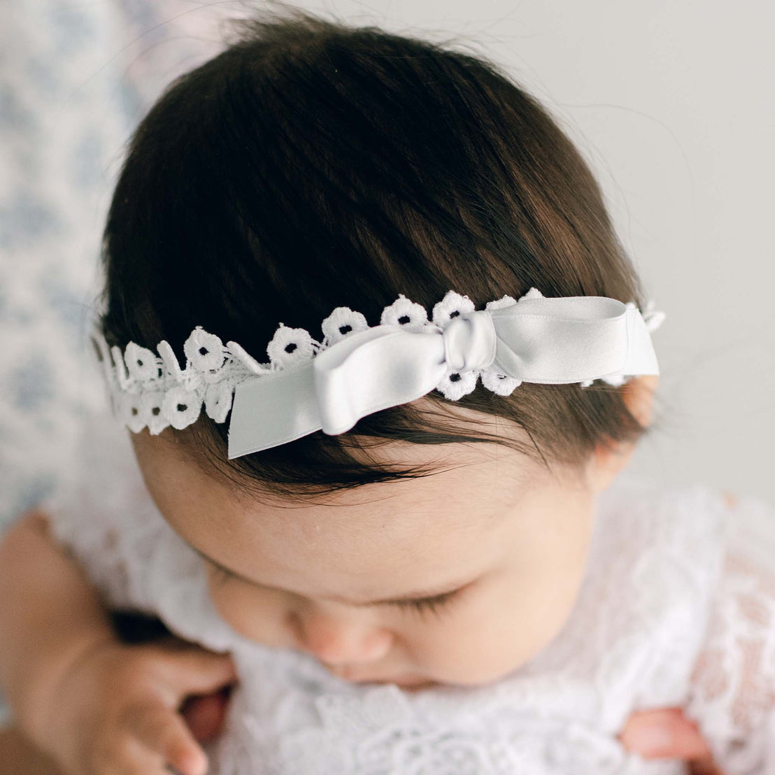 Close-up of a baby with dark hair wearing the Olivia Headband, looking downward. The background is softly blurred.