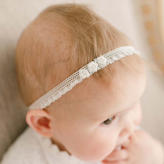 A close-up image of a baby with light hair, wearing the Eliza Lace Baby Headband decorated with small flowers, looking away from the camera.