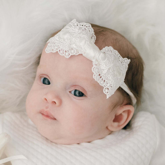 A close-up photo of a baby with bright blue eyes wearing a Madeline Lace Bow Headband, nestled in a soft white blanket.