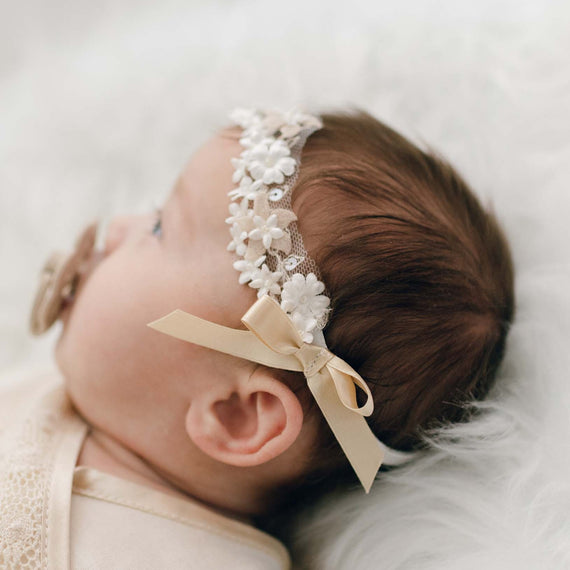 A newborn baby with dark hair wearing the Mia Headband with floral pearlesque beading and a gold silk ribbon bow, lying on a soft white blanket.