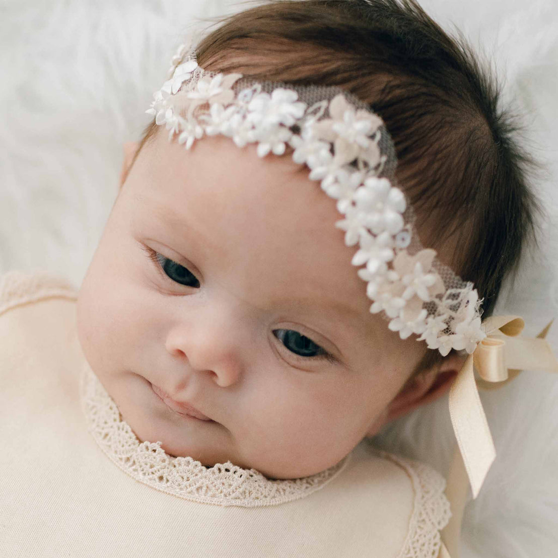 Close-up of a baby with blue eyes wearing the Mia Headband, featuring floral and pearlesque beading, along with the Mia Newborn Bib with lace details, lying on a soft white blanket.