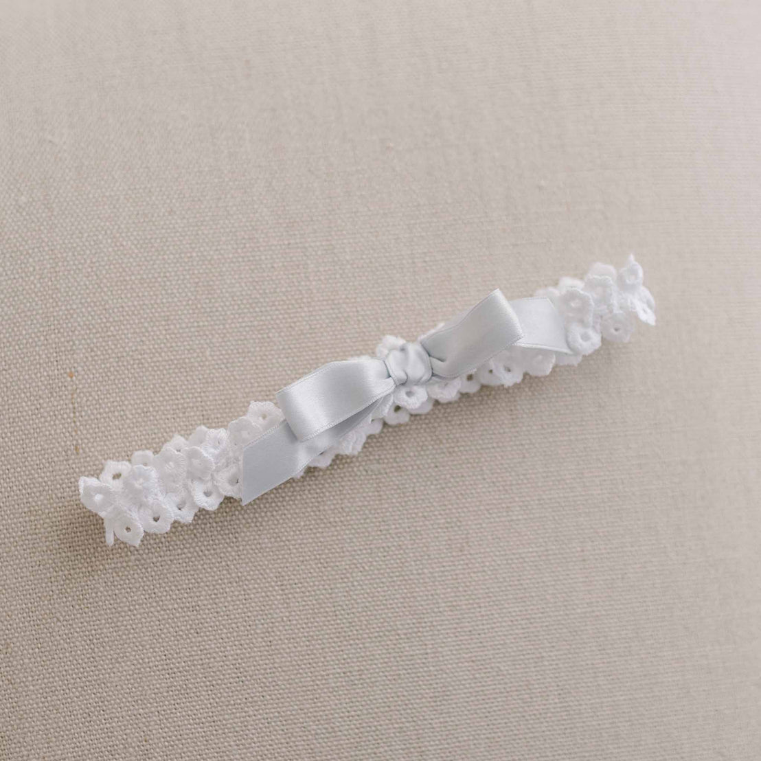 A delicate Olivia Headband made of white lace with floral patterns and a silky white ribbon tied in a bow, set against a neutral beige background, perfect for boutique baptism collections or as an heirloom piece.