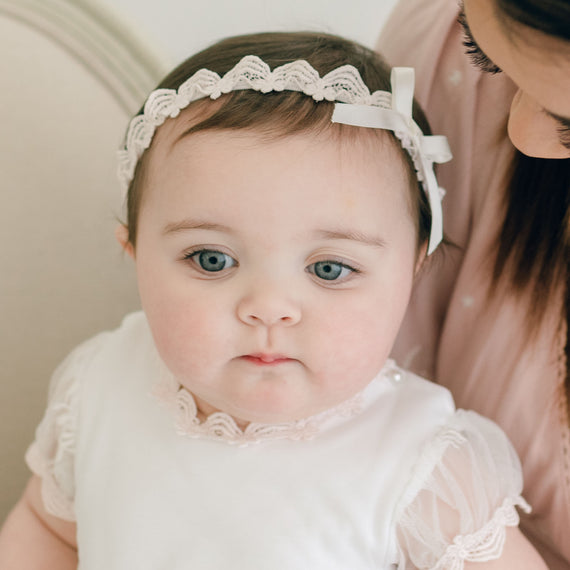 A baby with blue eyes and a Baby Beau & Belle Joli Lace Headband looks up, while a woman, partially shown, leans in close to the baby.