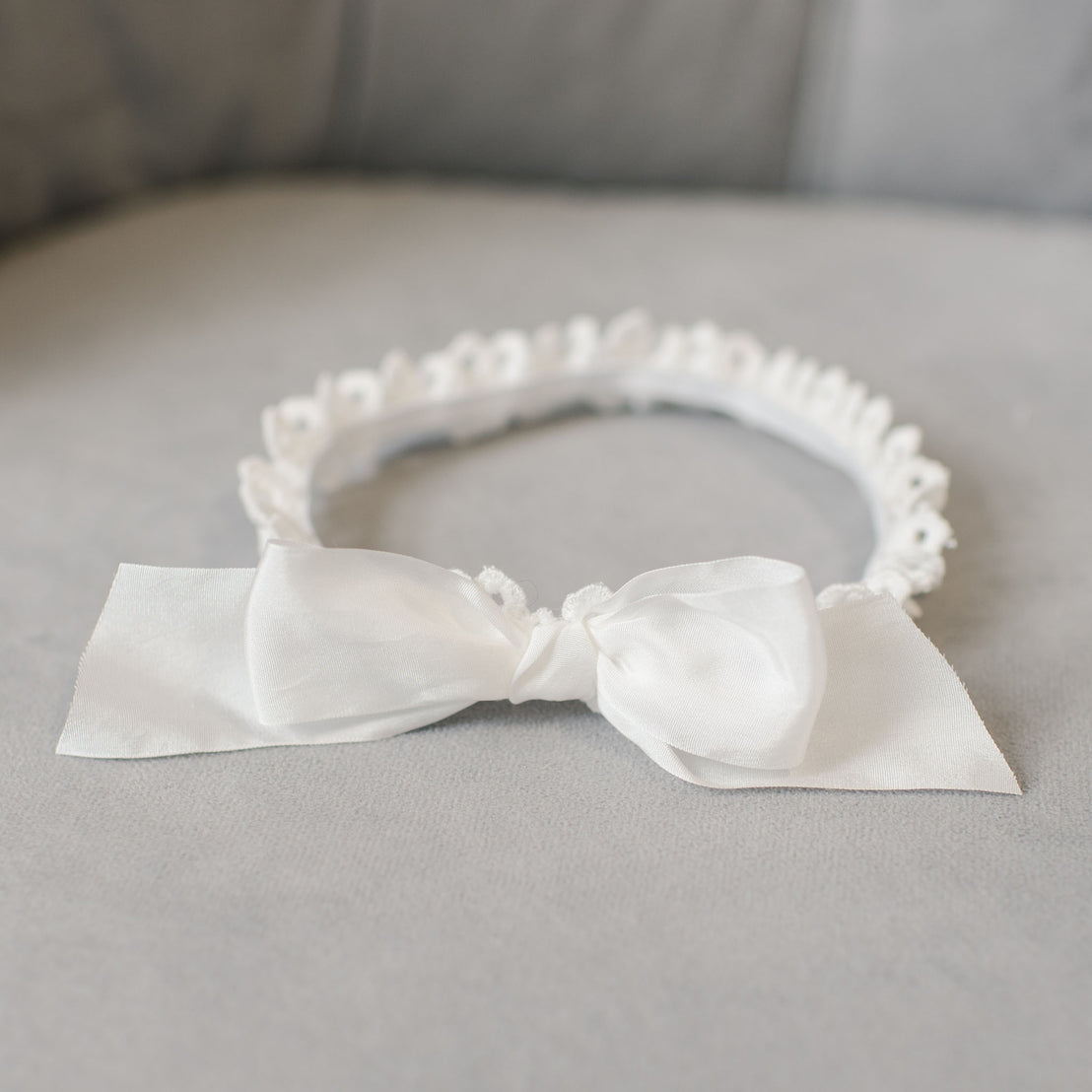 A delicate Grace Headband adorned with a large silk bow and small floral embellishments, perfect as a baby shower gift, resting on a soft gray cushioned background.