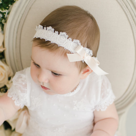 A baby girl wearing a white lace dress and the Melissa Headband, seen from above as she looks to the side. She is surrounded by soft, floral accents and an accessory bundle.