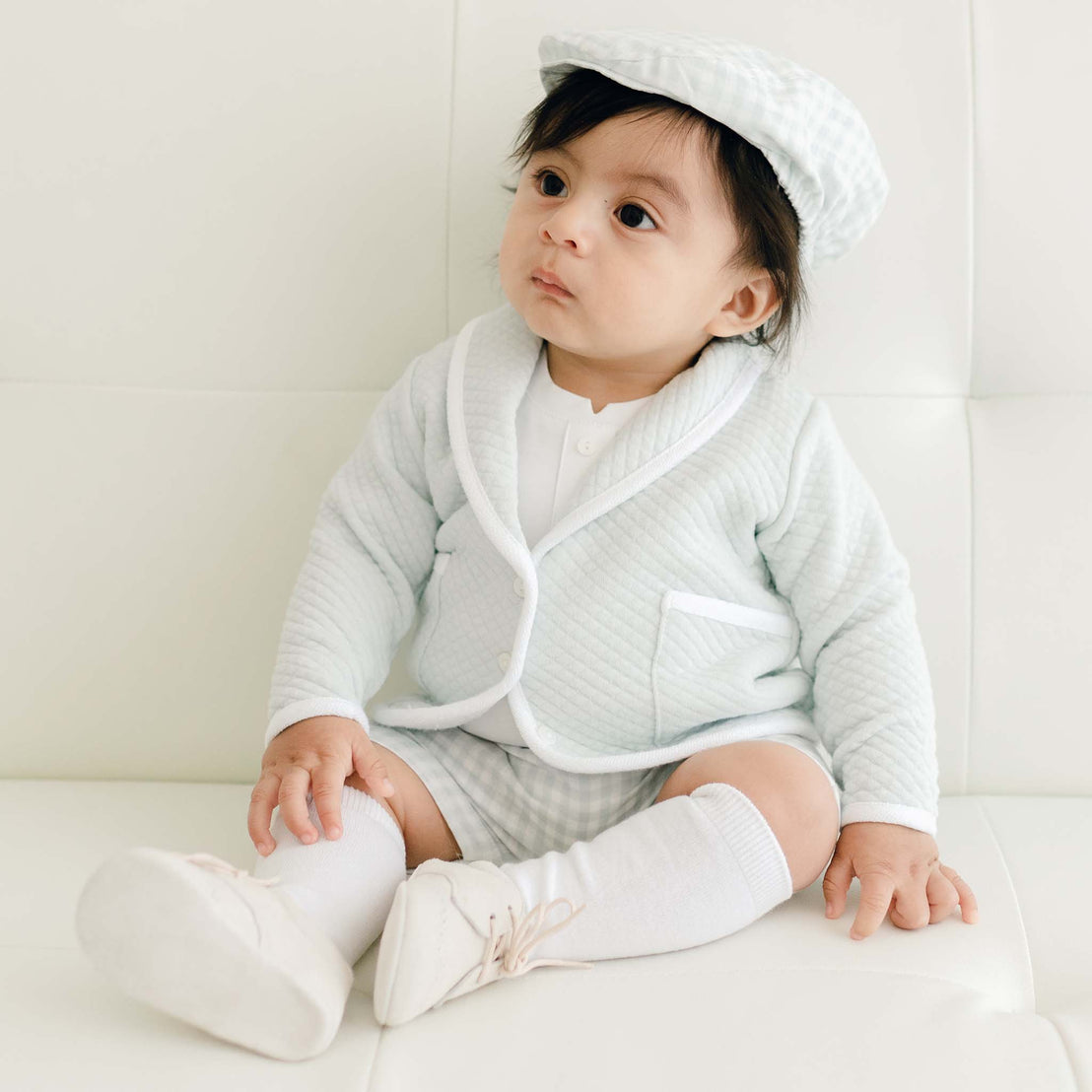 A baby in an Ian Cloud Shorts Set sits on a white couch, looking to the side with a curious expression.