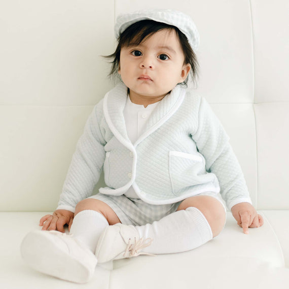 A baby dressed in an Ian Cloud Shorts Set sitting on a white couch, looking calmly at the camera with wide eyes. The baby wears a matching hat and white shoes.