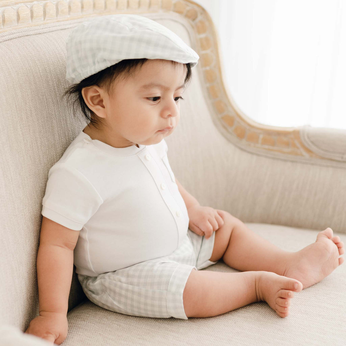 A baby wearing a Ian Cloud Shorts Set and traditional plaid shorts sits on a beige sofa, looking down thoughtfully while also donning a matching plaid cap.