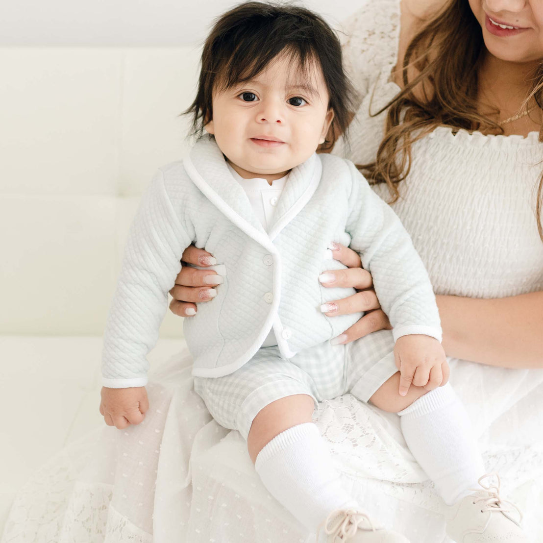 A baby with thick hair, dressed in an Ian Cloud Shorts Set and a designer white cardigan, sits on a woman's lap, both smiling in a brightly lit room.