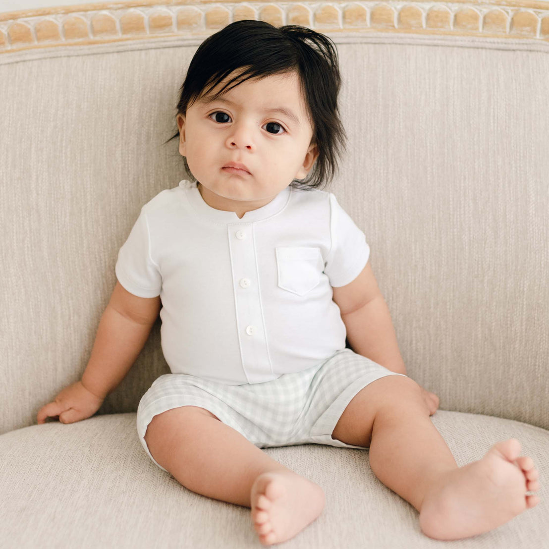 A baby with dark hair sits on a beige sofa, wearing the Ian Cloud Shorts Set, looking directly at the camera with a serious expression.