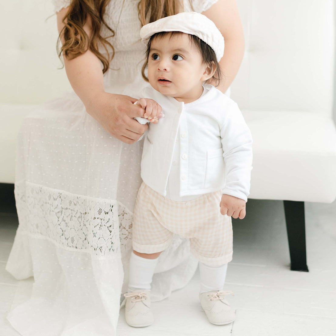 A toddler, wearing the Ian Fawn Shorts Set with a matching hat, stands holding a woman's hand, partially visible in a white dress, in a brightly lit room with white decor.