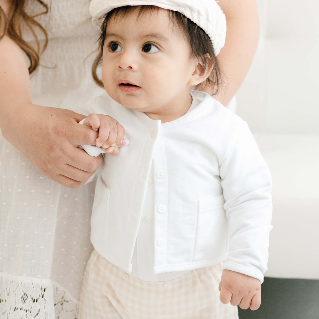 A mother holds the hand of her baby dressed in Ian Fawn Shorts Set. The baby, looking up with curiosity, has a white headband against a soft and light background.