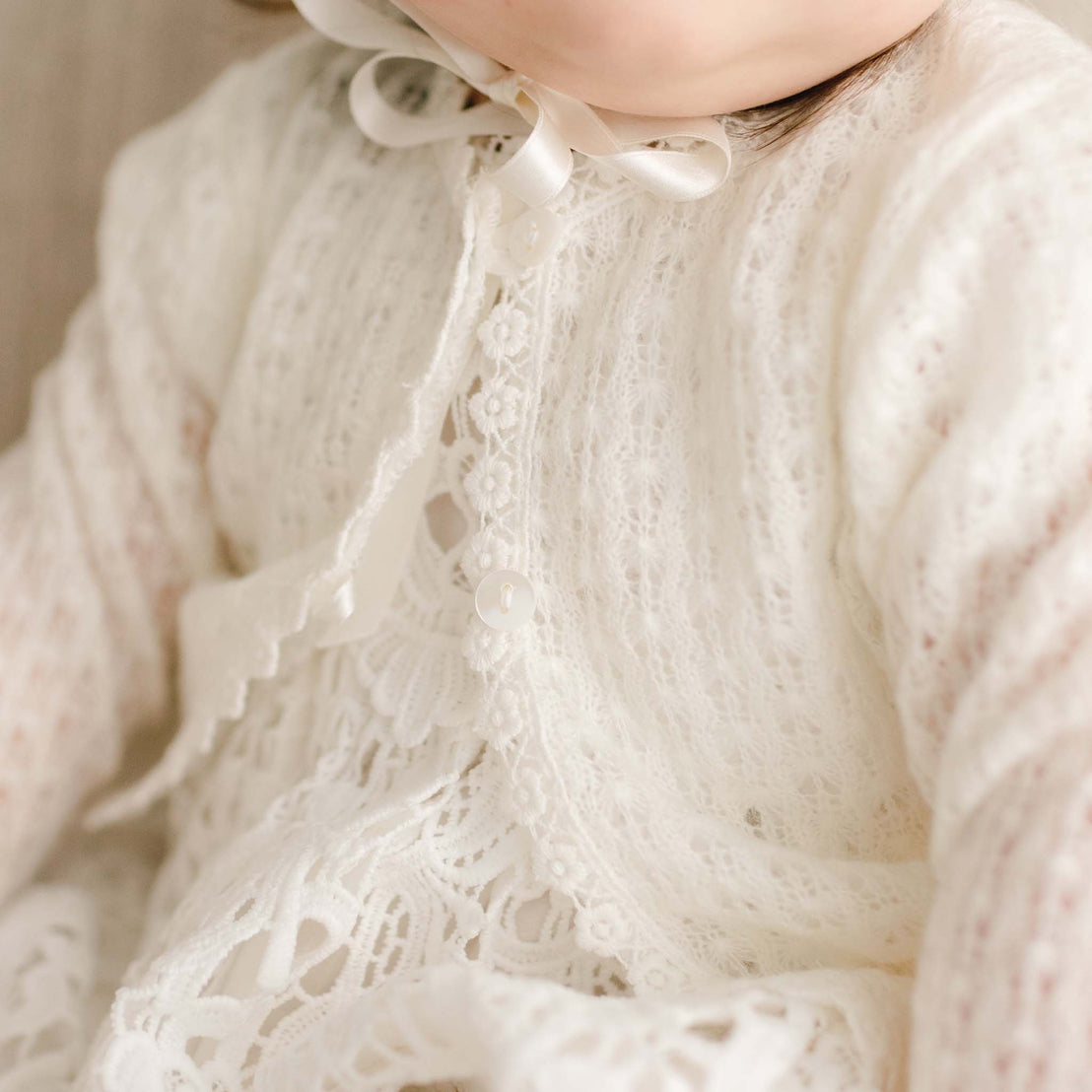 Detail of the Lola Knit Sweater. Baby girl wearing the Lola Knit Sweater that matches the Lola Christening Gown, Dress and Romper. Made of 100% ivory knit acrylic in light ivory, a soft and breathable knit with button closures and light ivory trim.