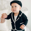 A baby wearing a black suit with white trim and a matching cap is holding an adult's hand. The baby looks upward with a neutral expression. The background is softly blurred, highlighting the baby.