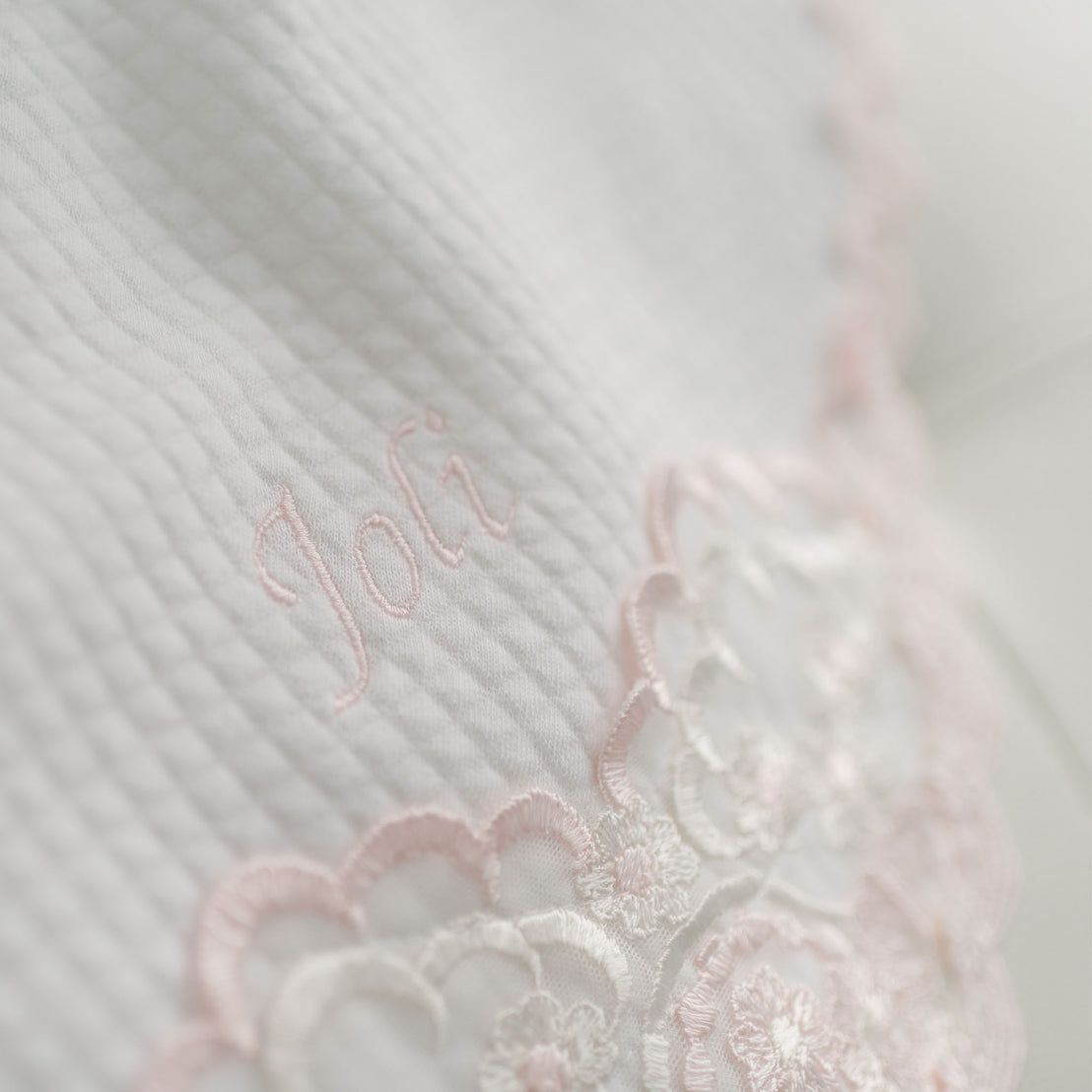 A close-up photo of a Joli Personalized Blanket with the embroidered name "Joli" in pink thread, partially overlaying an intricate white and pink lace design.