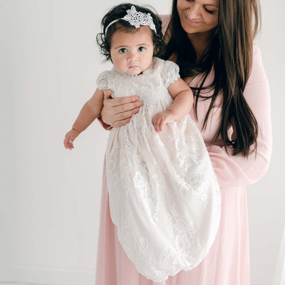 A woman in a pink dress holds a baby dressed in a Juliette Layette gown and a small headband, both looking towards the camera in a bright room.