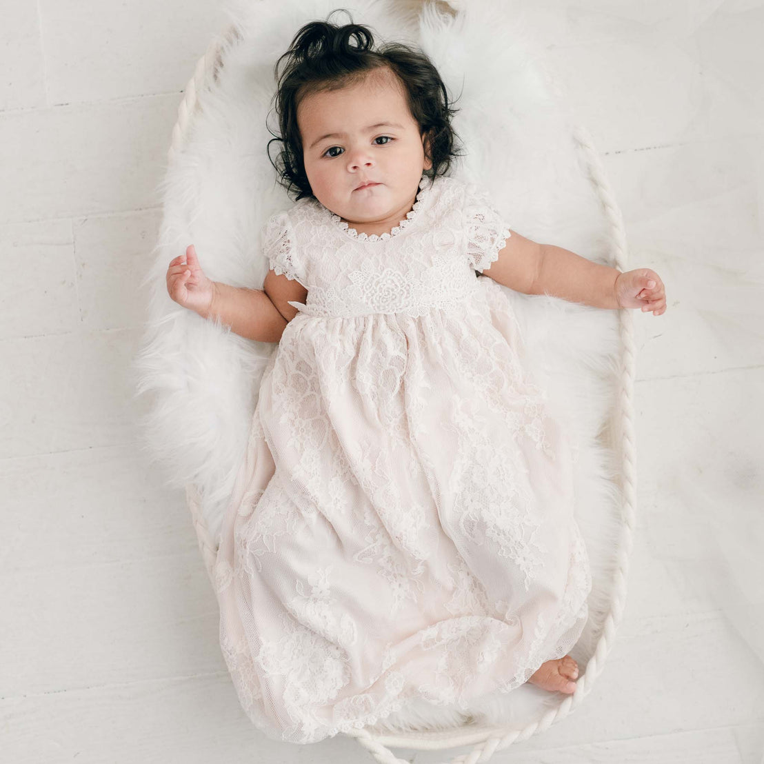 A baby wearing a white lace dress lies peacefully in a luxury, vintage-inspired Juliette Layette, viewed from above on a light background.