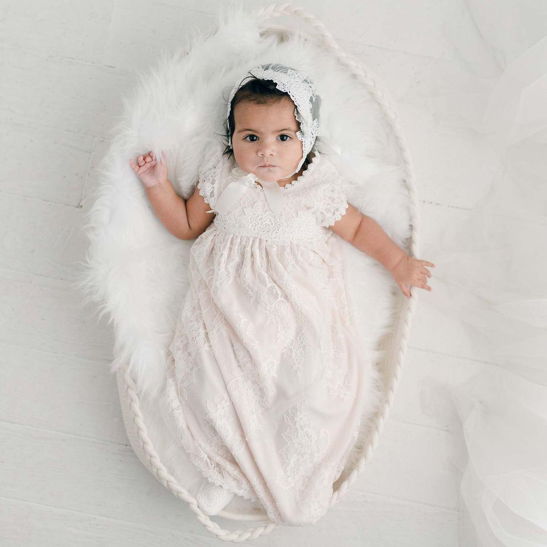 A baby wearing a Juliette Layette gown and headband lies in a handcrafted wicker basket adorned with white fur, looking directly at the camera with wide eyes.