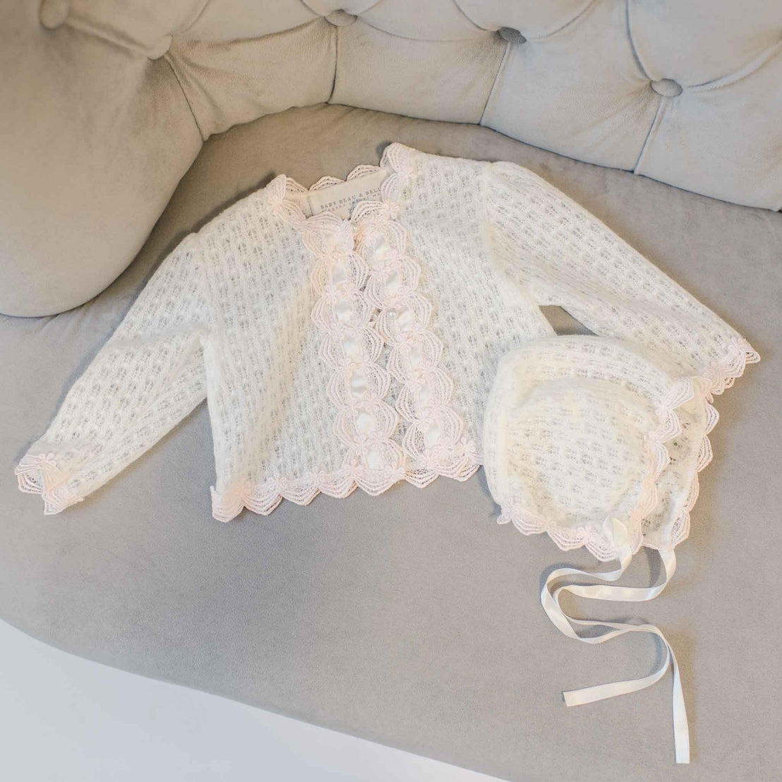 A delicate Joli Knit Sweater & Bonnet including a top, pants, and a bonnet, neatly laid out on a soft gray sofa.