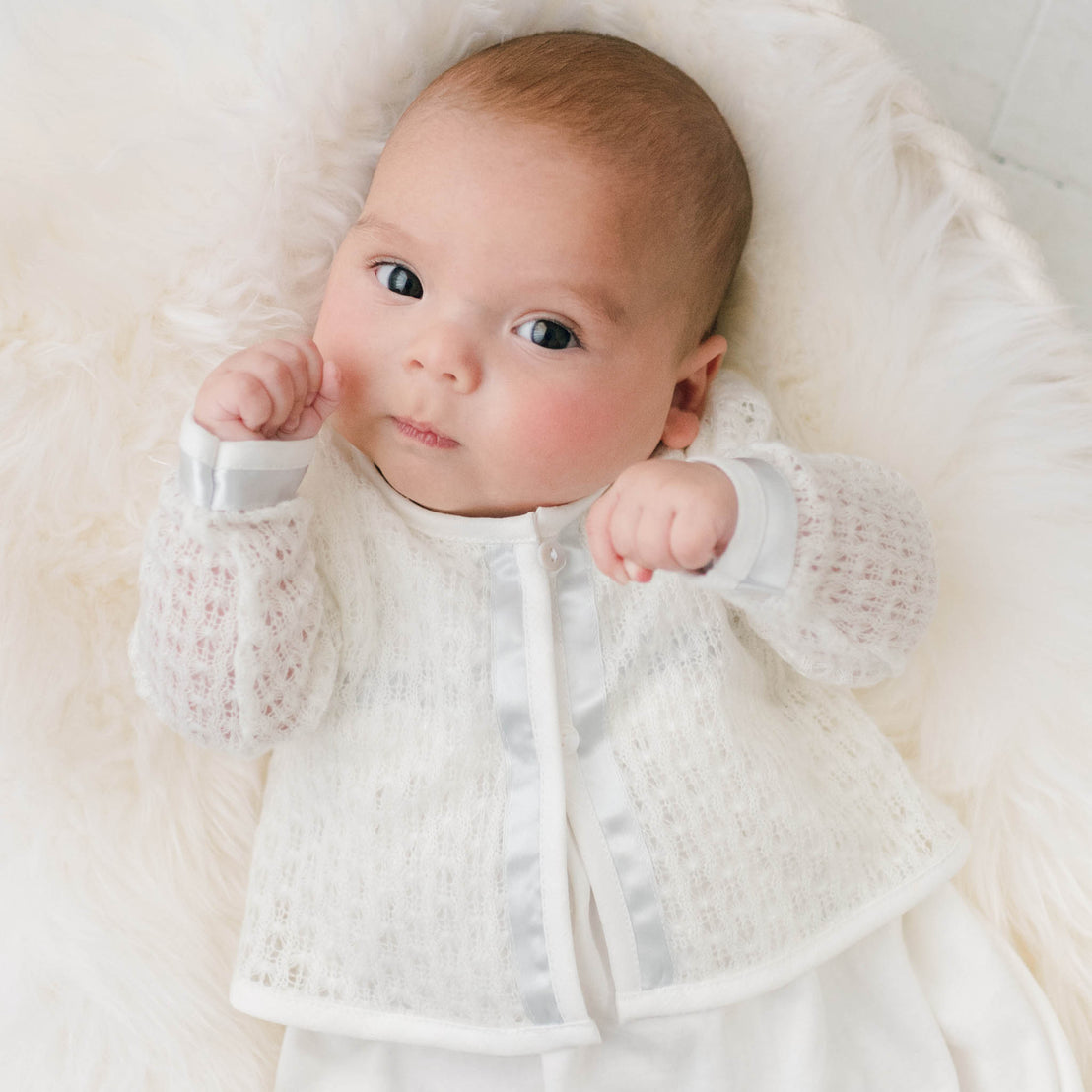 Newborn baby in a cradle and wearing the Owen Knit Sweater made from a soft knit in ivory and trimmed with ivory linen and blue silk ribbon