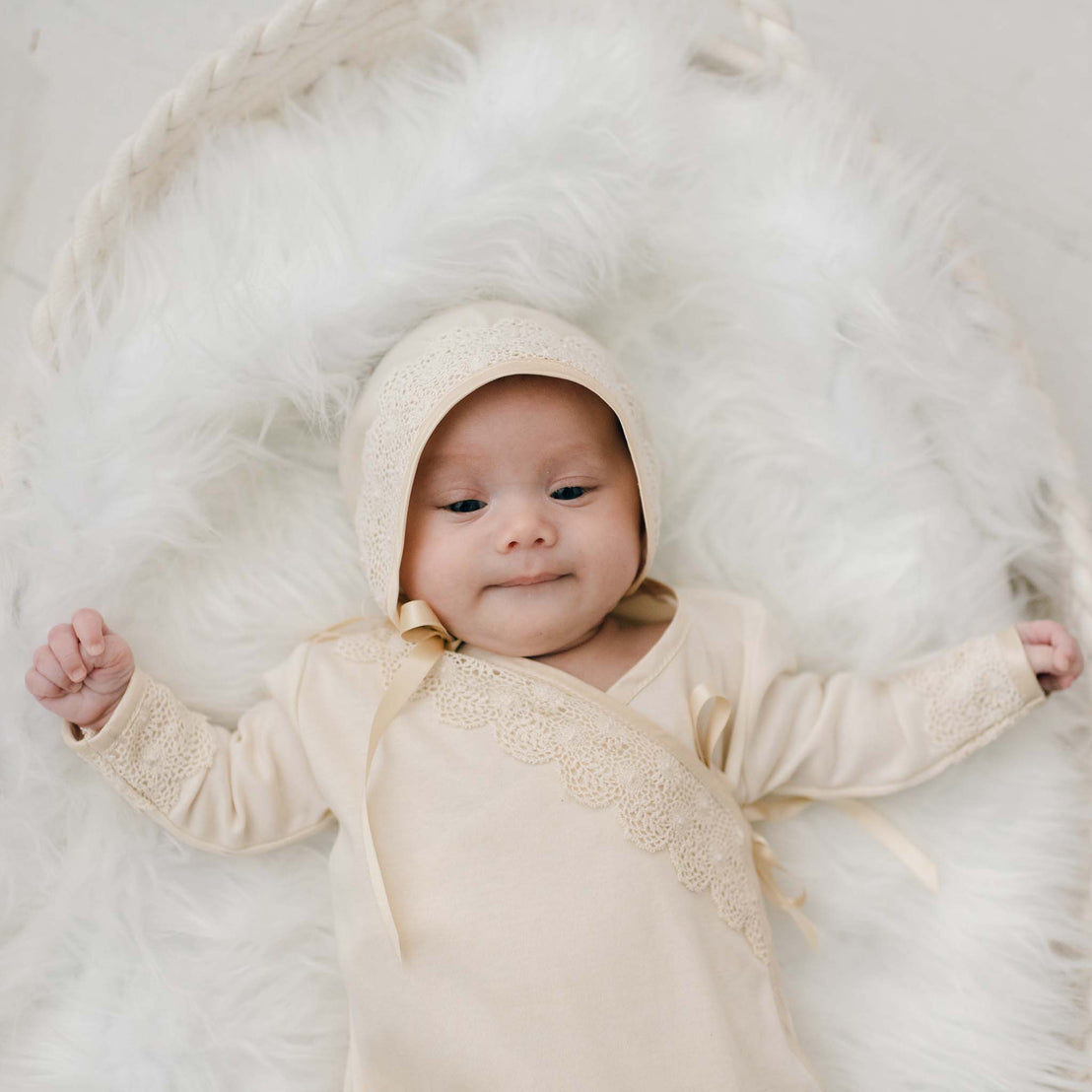 A serene newborn girl wearing the Mia Knot Gown & Bonnet lies on a white furry blanket, giving a gentle smile.