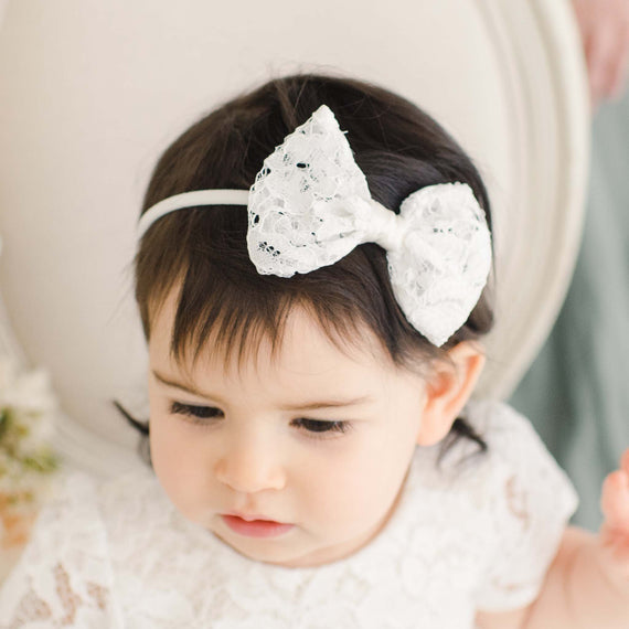 A close-up of a baby girl with dark hair dressed in a traditional white outfit for baptism, wearing a Rose Bow Headband and looking downward thoughtfully.
