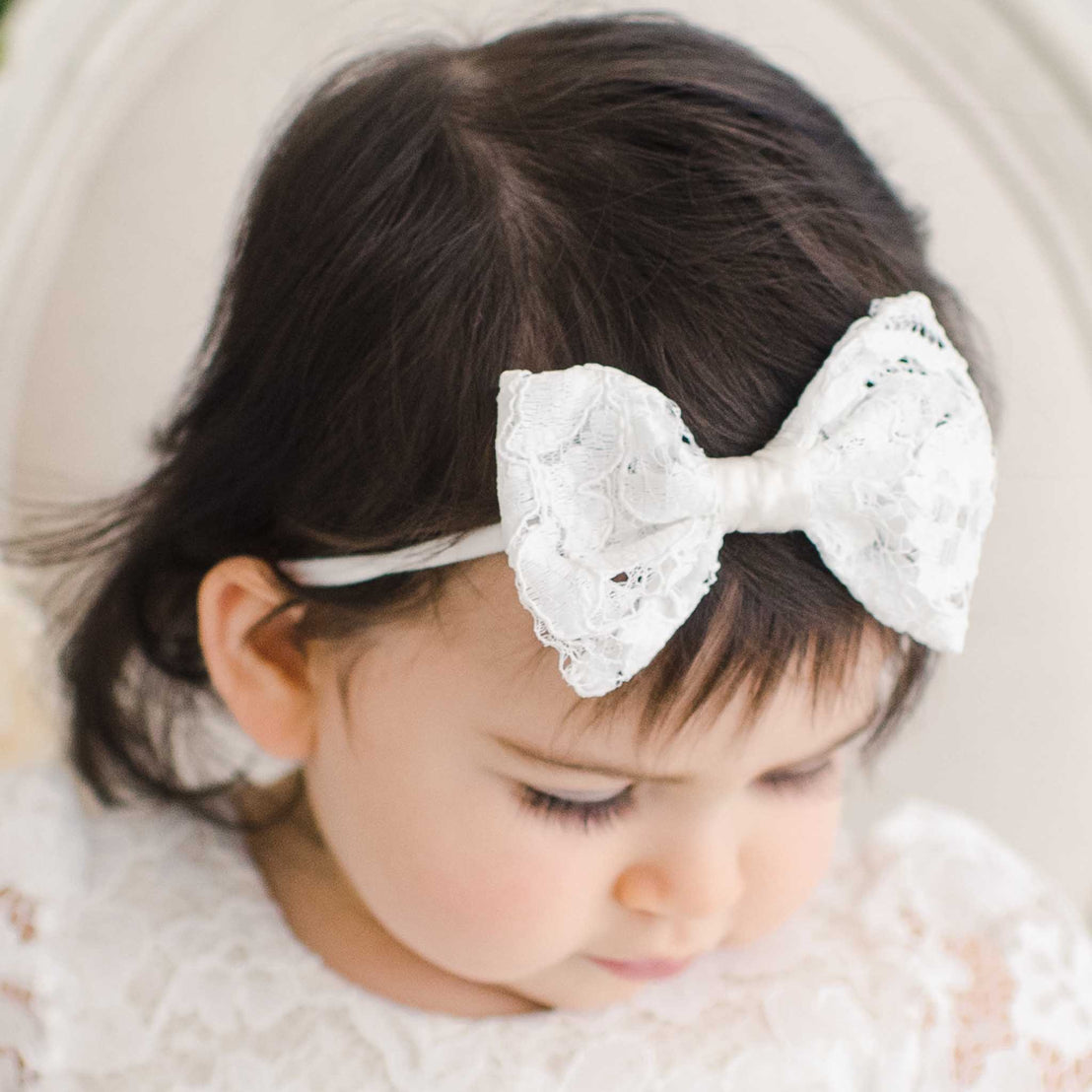 Close-up of a baby girl with dark hair wearing a Rose Headband, focusing on her gently closed eyes and detailed outfit for baptism.