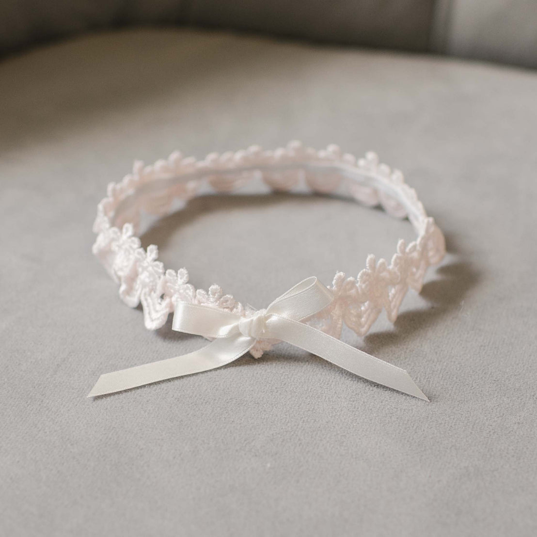 A delicate Baby Beau & Belle Joli Lace Headband adorned with a silk ribbon bow, displayed on a soft gray fabric background.