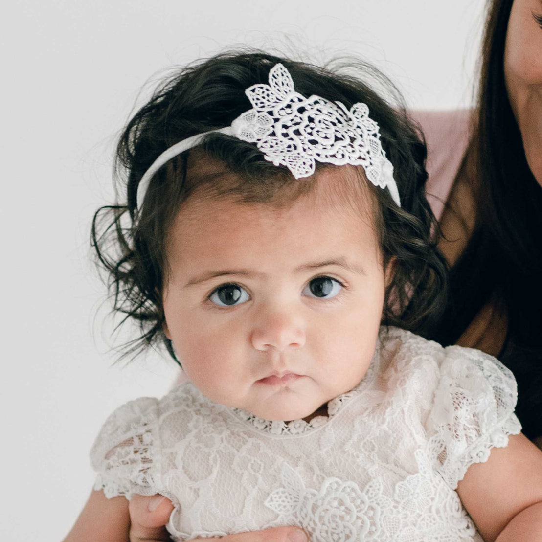 A baby with dark eyes and wavy hair wearing a white vintage lace dress and a white headband with a lace bow.