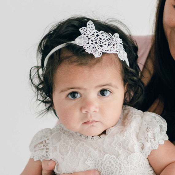 A portrait of a baby with large brown eyes and curly dark hair, wearing a handcrafted lacy white dress and a Juliette Lace Headband with a lace bow, looking directly at the camera.