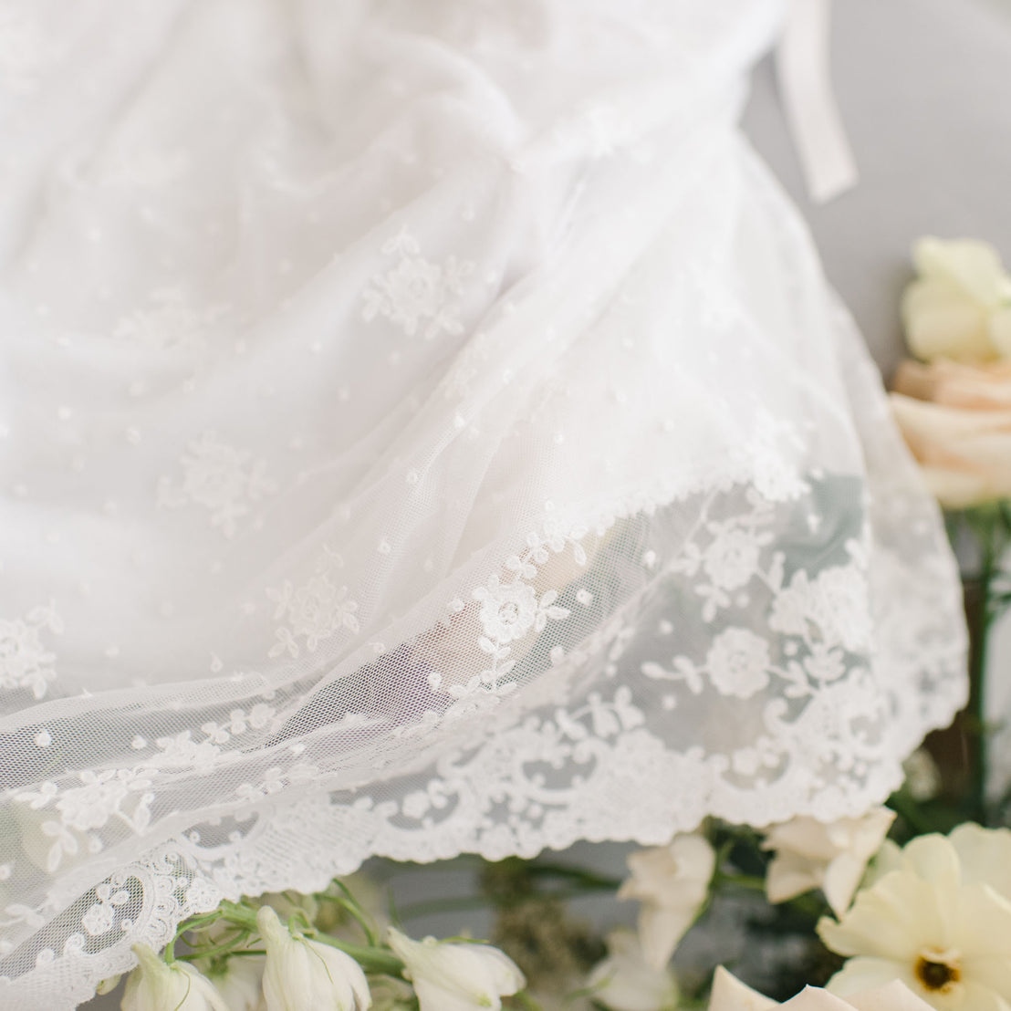 Close-up of a delicate white Melissa Christening Gown & Bonnet featuring detailed lace patterns, partially draped over soft pink and white flowers.