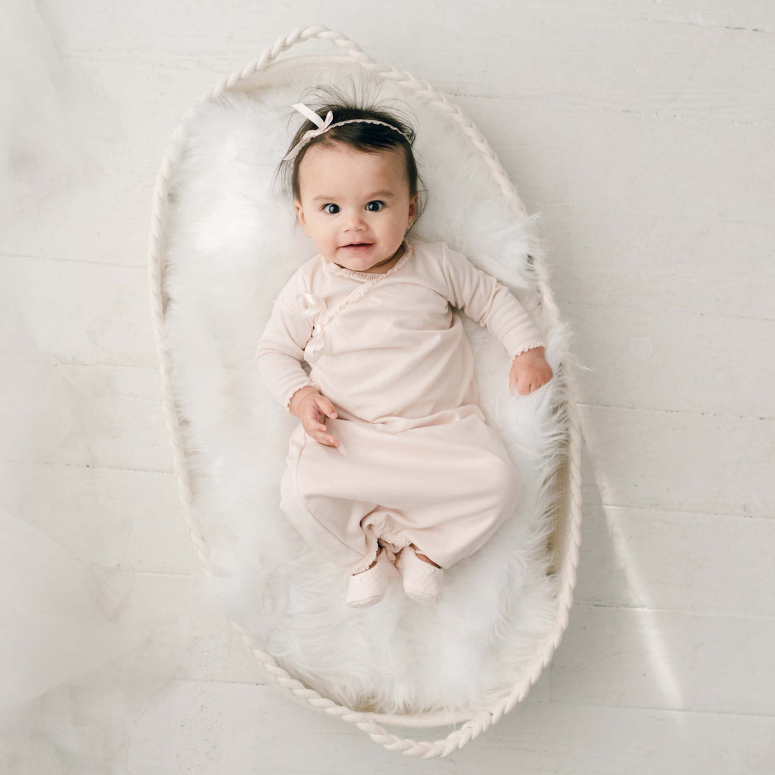 A cute newborn girl with dark hair wears the Ava Headband and matching Ava Newborn Layette and Ava Quilted Booties lies in an oval-shaped basket lined with soft white fur and looks upwards with a slight smile