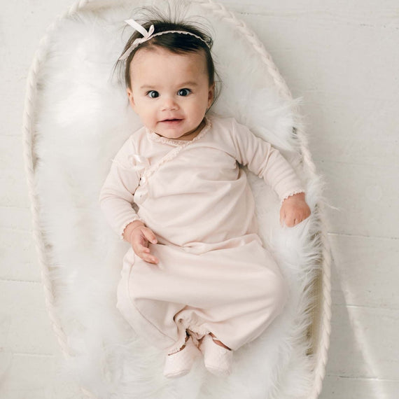 A baby wearing the wearing the Ava Newborn Layette & Headband, a pima cotton layette in a shade of blush pink and matching blush ribbon bow headband, lies smiling in a white fluffy blanket.
