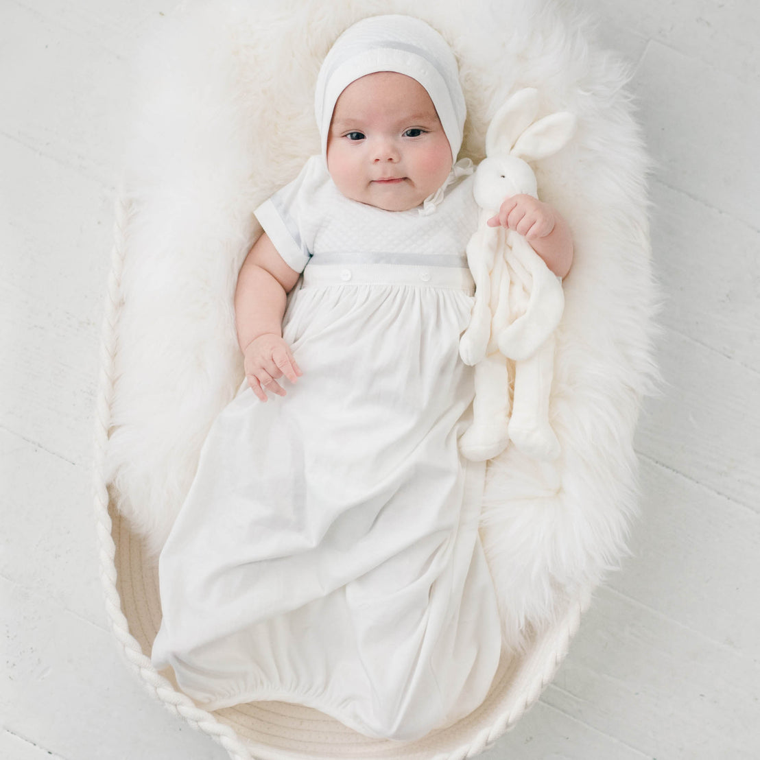 Newborn baby boy sitting in a white fur-lined crib. He is wearing the Owen Layette and Quilted Newborn Bonnet. He is also holding the Owen Silly Bunny Buddy