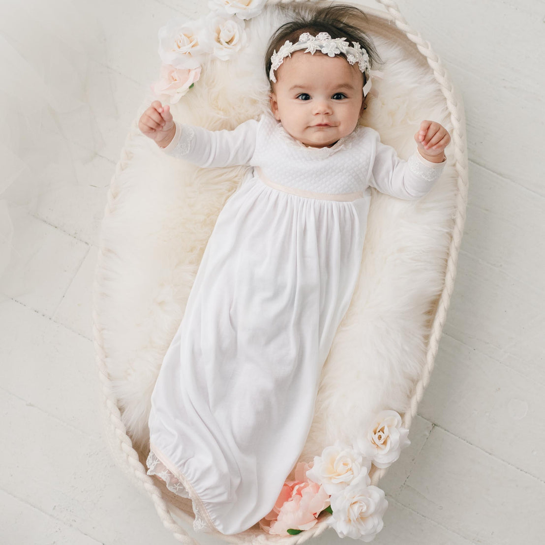 Baby girl wearing the Tessa Quilt Newborn Gown and laying in a crib. The newborn gown is made with soft pima cotton in white and features a plush white quilt bodice with an ivory Venice lace accent along the neck and cuffs.