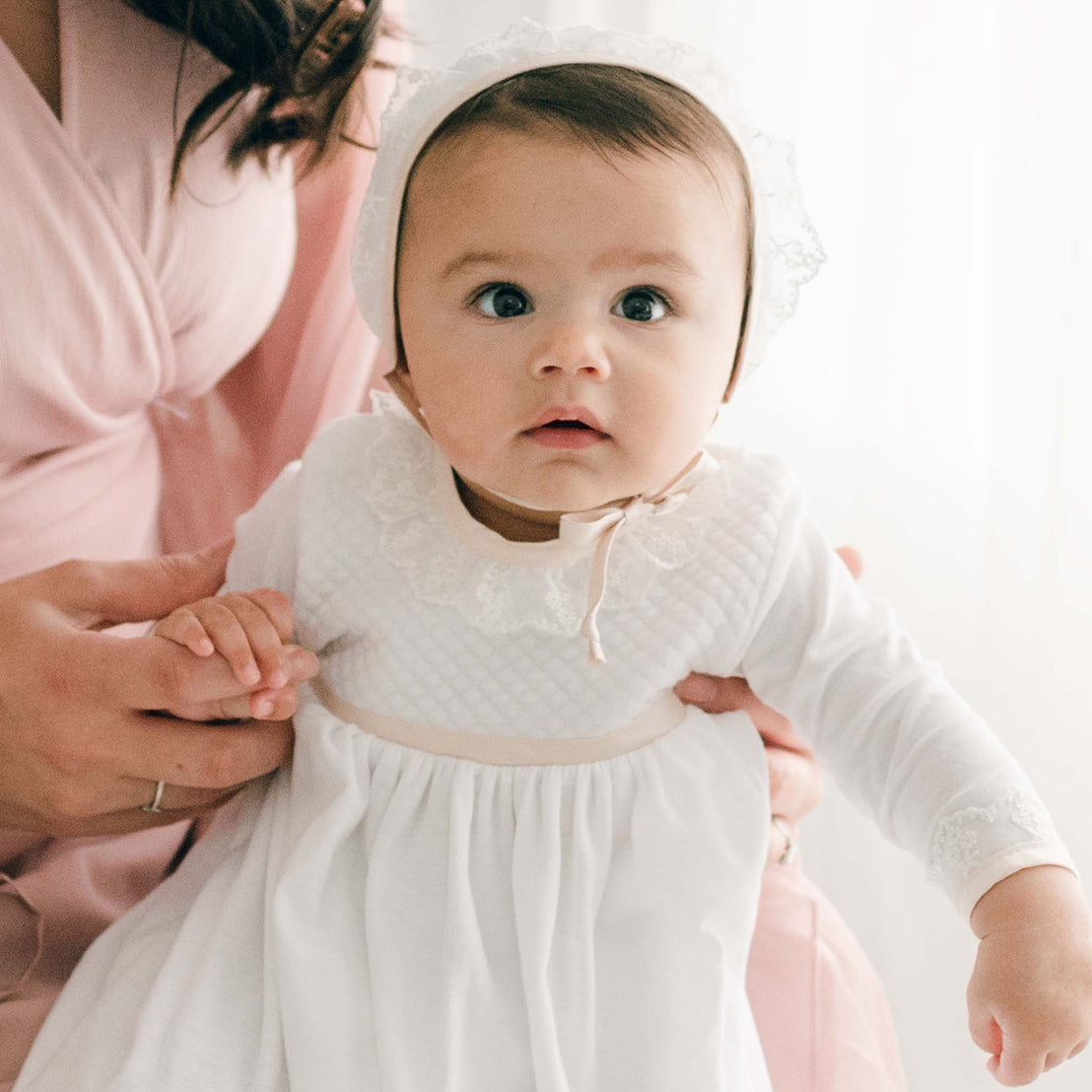 Close up photo of a newborn baby girl wearing the Tessa Quilt Newborn Gown accompanied by her mother. The newborn gown is made with soft pima cotton in white and features a plush white quilt bodice with an ivory Venice lace accent along the neck and cuffs.