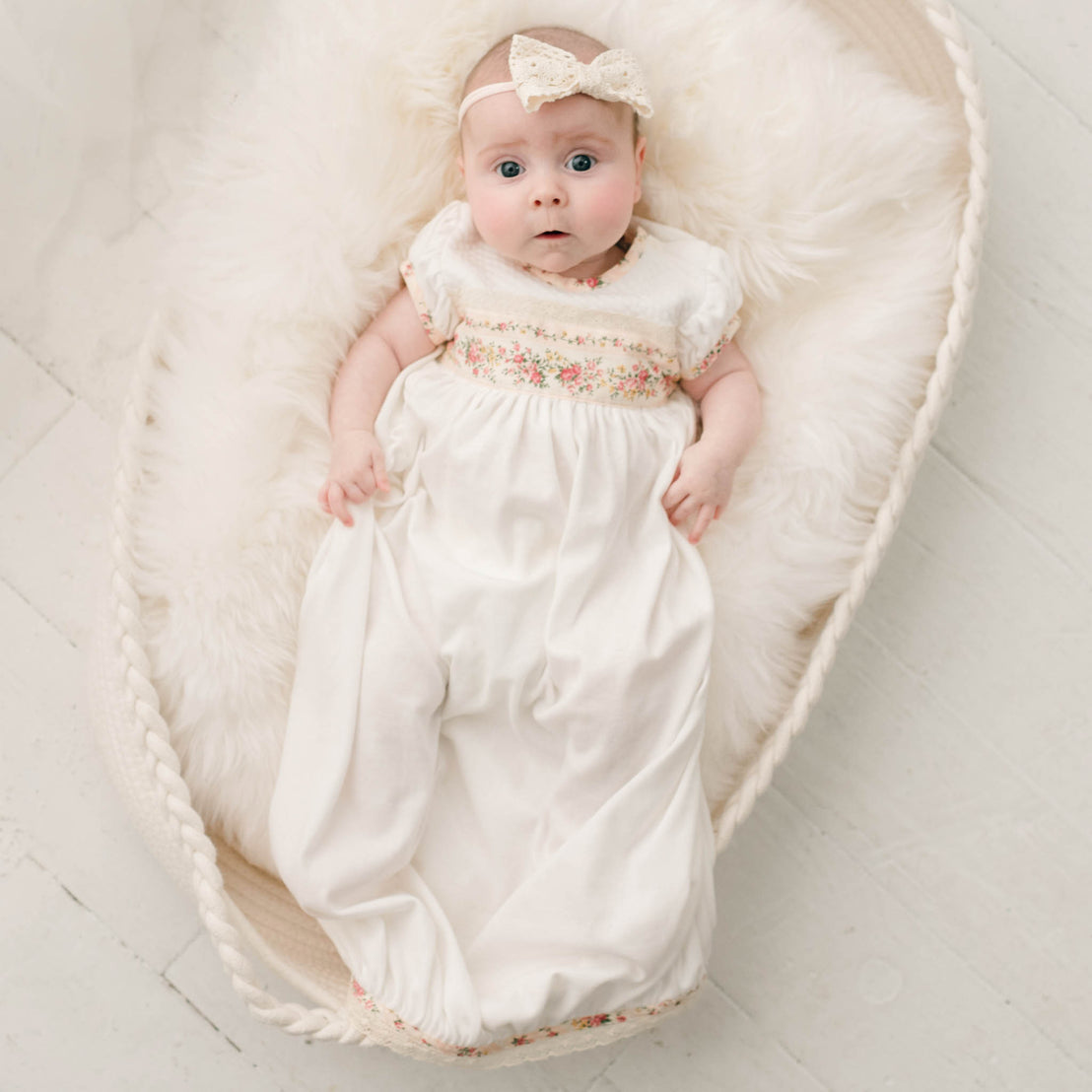 Newborn baby girl in a crib and wearing the "Blush" Eloise Layette Gown with matching Eloise Headband.
