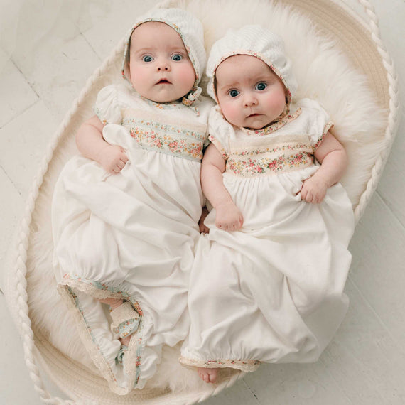 Two twin baby girls wearing the Eloise baby layette gown-one in "Powder Blue" and the other in a "Blush" floral pattern. They are also wearing the matching Eloise Quilted Bonnet.