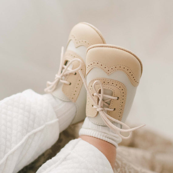 Baby wearing a pair of the Beige and Ivory Wingtip Shoes made with a tan and ivory matte leather with tan suede soles and detailed edging.