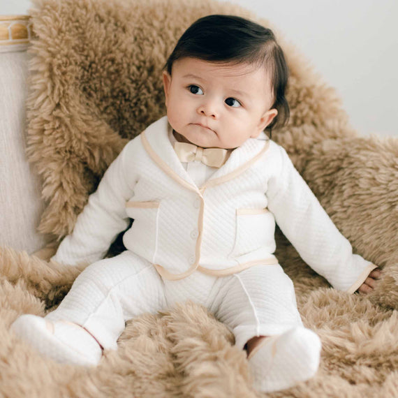 Baby sitting on a chair with brown fur. He is wearing the Liam 3-Piece Suit made with soft ivory quilted cotton and champagne silk trim. He is also wearing a champagne silk bowtie 