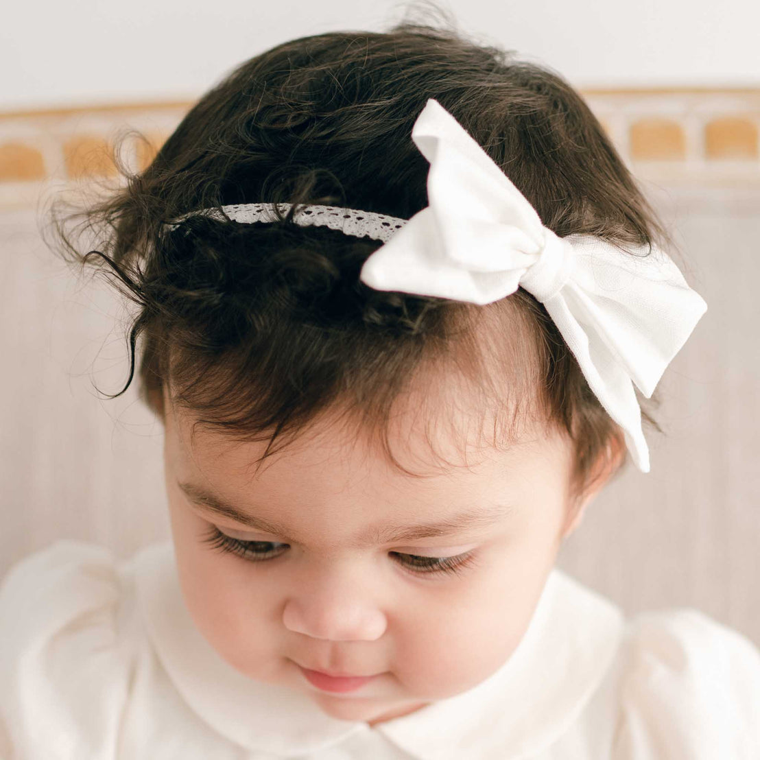 A close-up portrait of a toddler with curly hair, dressed in a white boutique outfit for a baptism, looking down thoughtfully wearing the Emma Linen Bow Headband.